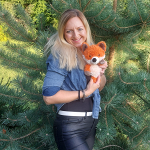 Maria hält Wurzelfuchs - ein flauschiger Stofftierfuchs - im Arm vor einem Nadelbaum